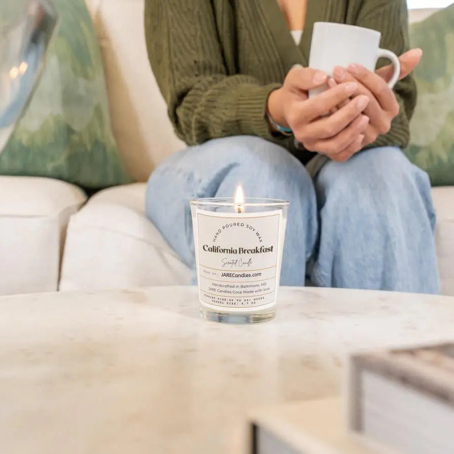 Person holding mug with California Breakfast JARE soy candle lit in cozy living room, orange pineapple scent
